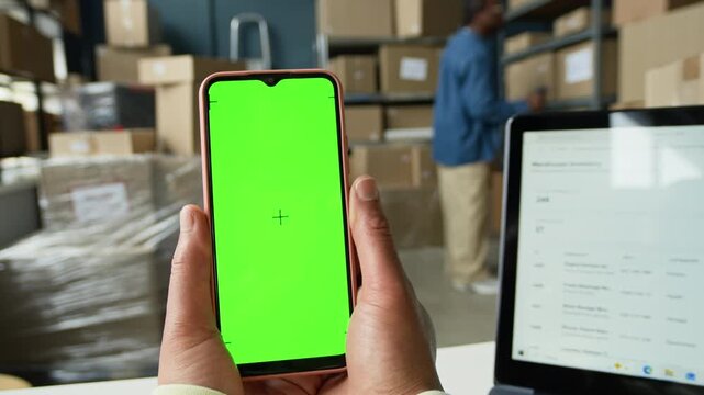 Worker holding smartphone with green screen in warehouse with African American colleague managing inventory. Perfect for promoting logistics software or delivery tracking apps