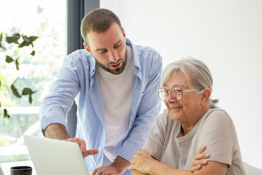 Young Man Helping Senior Woman With Laptop At Home While Teaching Technology Skills