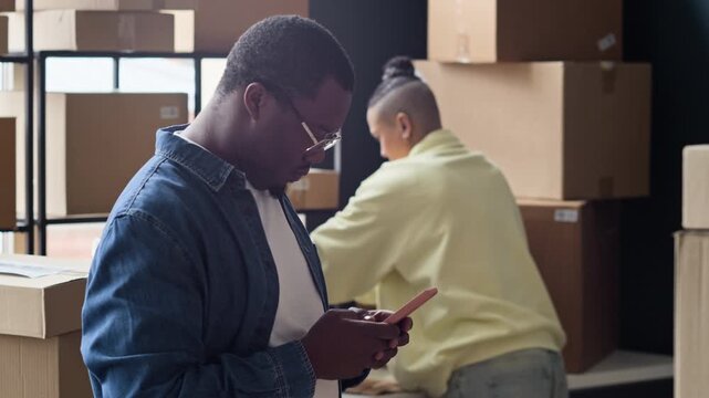 Medium shot of African American young man using smartphone in warehouse while colleague organizing cardboard boxes. Footage perfect for e-commerce logistics and order fulfillment software commercials