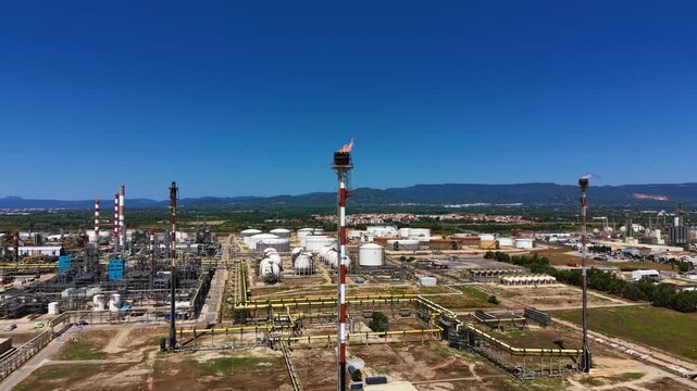 Aerial view of an active gas flare stack burning above a large petrochemical refinery in Spain, featuring yellow pipelines, spherical LPG vessels, storage tanks, and industrial chimneys