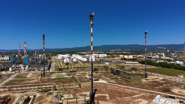 Aerial view of a large petrochemical refinery in Spain featuring an active gas flare stack, yellow pipeline network, storage tanks and chimneys under a deep blue sky with mountains