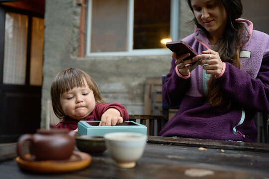 Woman Sits With Child at Table While Using Smartphone Outside
