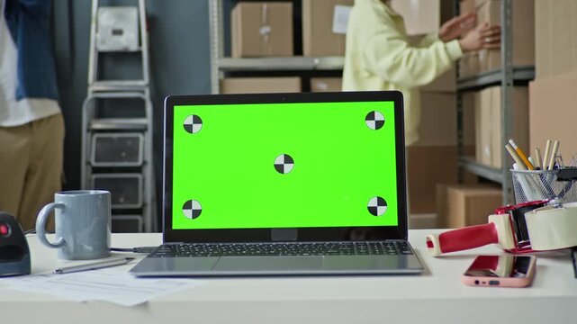 Focus on laptop with green screen displaying chroma key sitting on desk in warehouse while workers manage inventory in background. Perfect for showcasing e-commerce software or logistics apps