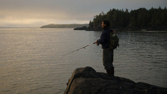 Fisherman stands on rocks by the water during sunset