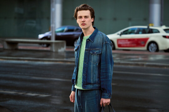 Young Man Stands on City Street in Denim Jacket With Bag