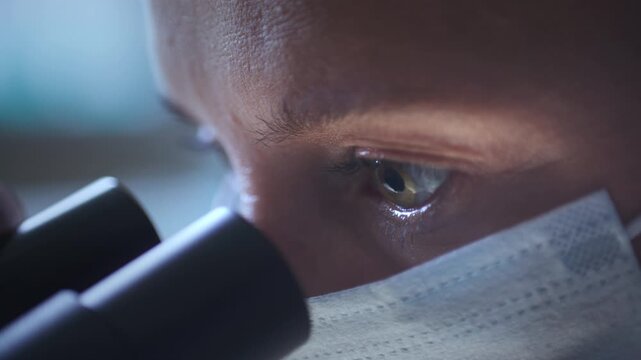 Macro shot of eyes of focused woman scientist in protective face mask examining medical specimen through microscope in laboratory