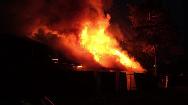A house fire causes bright flames and heavy smoke at night, illuminating the dark surroundings and creating a chaotic scene.