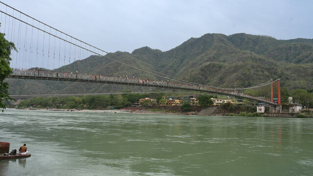 Pedestrians (from distnace) crossing the iconic Ram Jhula suspension bridge over the green waters of the Ganges river, Rishikesh, Uttarakhand, India.