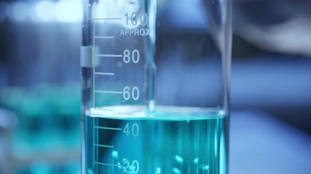 Glass beaker with graduated measurement markings being filled with blue chemical solution during scientific research or medical test in laboratory. Close-up, tilt-up shot