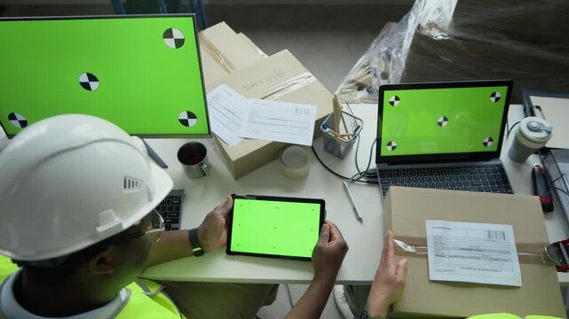 View from above of young African American male warehouse manager using green screen tablet while colleague packs cardboard box near laptop. Ideal for logistics software promos and e-commerce ads