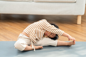 Fototapeta premium Asian preschool girl holds seated side stretch on yoga mat with arm overhead and leg extended, playful home workout moment, healthy flexibility practice and child wellness routine at home indoors.