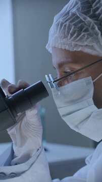 Professional medical researcher wearing face mask, disposable cap and protective gloves analyzing samples with microscope in clinical laboratory. Vertical shot, side profile