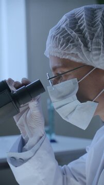 Female scientist in protective face mask, hairnet, gloves and lab coat looking into microscope eyepiece during clinical research or laboratory testing. Vertical clip, side view