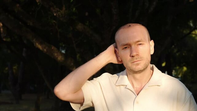A portrait of a thoughtful middle-aged man after a hair transplant operation, scratching abd rubbing his itchy shaved head with a worried look, standing outdoors in Portugal during golden hour.
