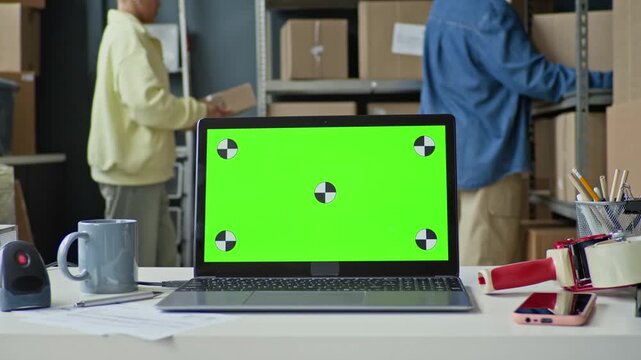 Focus on laptop with green screen on desk, multiethnic warehouse workers organizing boxes in background. Perfect for showcasing logistics software or e-commerce tools