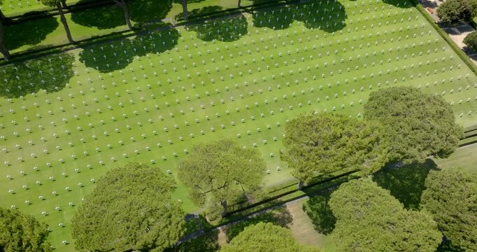 Aerial view of a military cemetery with rows of white headstones on a lush green lawn, surrounded by green pine trees and shadows. It is a beautiful sunny morning.