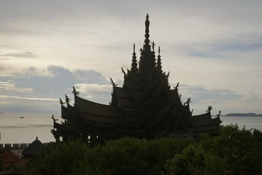 scenic silhouette of a grand multi-tiered traditional structure of 'sanctuary of truth' with ornate spires rising behind lush green treetops overlooking the calm sea under an overcast sky