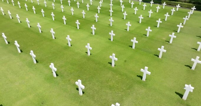 High angle view of rows of white marble crosses standing in perfect alignment on a pristine green lawn, casting shadows under bright sunlight inside an american military cemetery.