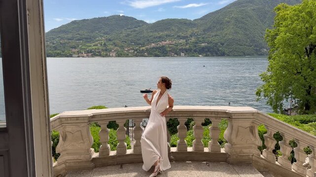 Beautiful Elegant Woman in White Halter Dress and Sunglasses on the Baroque Stone Terrace of a Historic Italian Villa With a Breathtaking Panoramic View of Lake Como and Alpine Mountains, Lombardy Ita