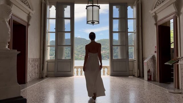 Elegant Young Woman in Backless White Dress Walking Toward the Grand Doorway of a Historic Italian Villa Overlooking Lake Como and Alpine Mountains, Lombardy Italy
