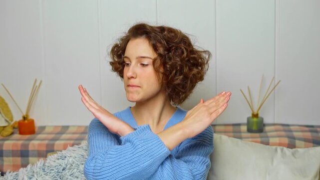 young woman with curly hair showing a stop gesture with crossed arms, expressing denial or refusal while biting her lip in a cozy indoor setting