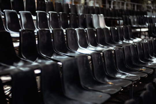 Rows of vacant black plastic chairs in dark spectator stands, with shallow focus and a few white seats creating contrast for venue, event and audience themes