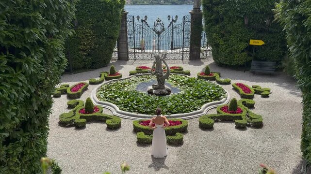 Beautiful Young Woman in White Dress Walking Through a Historic Italian Villa Garden Toward an Ornate Wrought Iron Gate Revealing Lake Como, Lombardy Italy