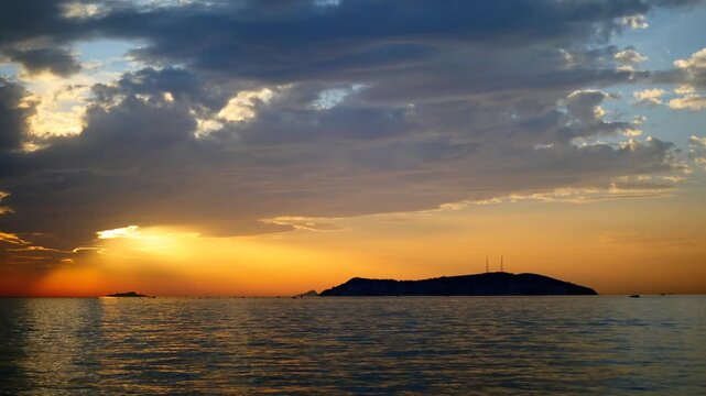 Dramatic sunset over the Sea of Marmara, with the sun breaking through thick clouds to cast a golden orange glow on the water surface near prominent silhouette of Kinaliada Island in Istanbul, Turkey
