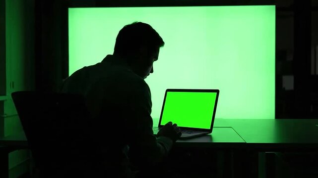Man works on laptop in dark room with green screen light