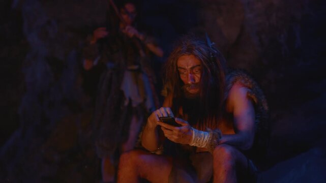 A caveman using modern technology while being situated in a distinctly primitive setting