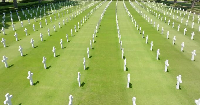 Aerial view of symmetric rows of white marble crosses on a green lawn at an American military cemetery, honoring fallen soldiers. It is a summer sunny morning. The cemetery is empty.