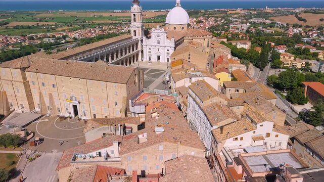 Loreto Marche Italy 4K Aerial. Drone fly through between basilica dome and bell tower with camera tilt up over historic square and sea background. Ancient Roman Catholic sanctuary pilgrimage 