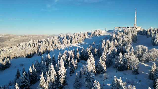 Aerial view of the snow-covered slopes and pine forests of Col de la Faucille with a radio tower on the mountain peak in Gex, Auvergne-Rhone-Alpes, France.