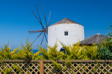 Whitewashed Santorini windmill with wooden sails under clear blue sky, framed by lush shrubs and lattice fence. © Andrei Tomas