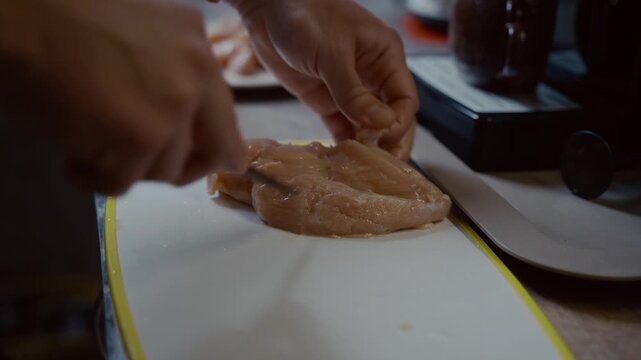 Close-up hands of Caucasian young male are cutting fresh chicken meat on plastic white board at home in kitchen. Hobby cooking, bachelor making dinner. A lonely man prepares poultry for lunch.