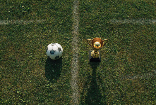 A classic black and white soccer ball and a shiny gold trophy cup standing side by side on the green grass of a soccer field pitch surface