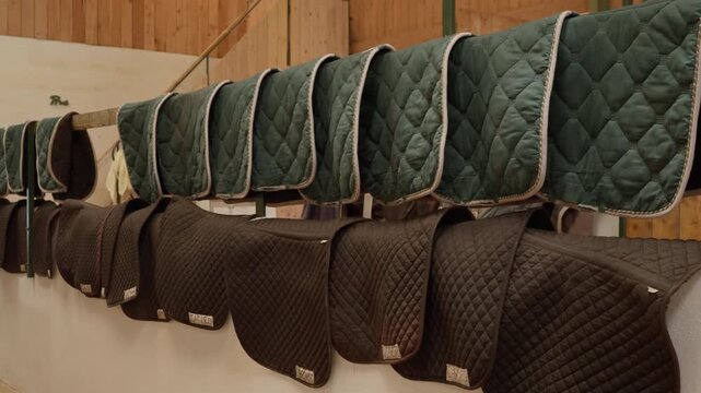 Equestrian saddle pads hanging on storage racks inside a professional stable. Rows of quilted horse blankets and riding equipment for equestrian sports training on a countryside ranch farm.