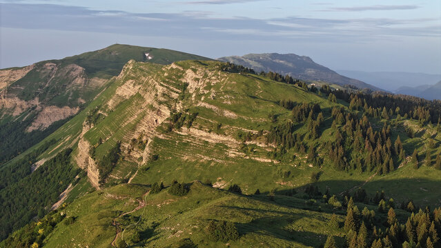Aerial view of Col de la Faucille mountain pass with lush green slopes, rocky ridges, and coniferous forests under soft morning light Gex, Auvergne-Rhone-Alpes, France.