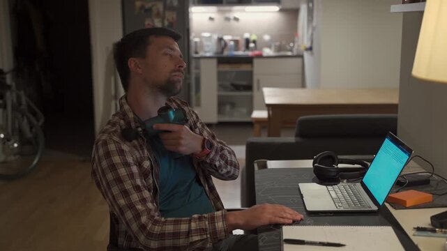Neck pain, cervical osteochondrosis. Male student studying until night at computer with neck pain. Tired guy kneads neck with pistol shock massager while working on laptop. Muscle recovery and massage