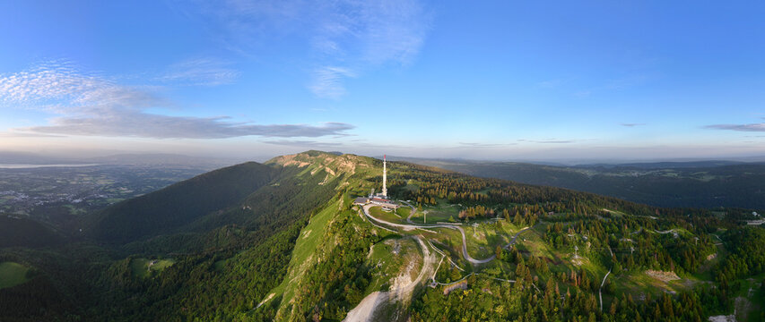 Aerial view of Col de la Faucille featuring a prominent communications tower, lush green mountain slopes, dense pine forests, and winding roads in Gex, Auvergne-Rhone-Alpes, France.