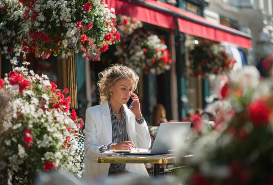 A woman in a white blazer sitting at an outdoor cafe table, talking on her mobile phone with a laptop computer open in front of her, flowers