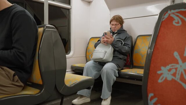 Senior woman puts on eyeglasses and uses smartphone inside Paris commuter train carriage. Elderly female passenger types on mobile phone screen, browsing online internet connection and smiling.
