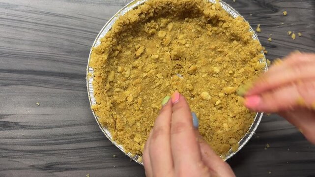  woman presses biscuit crumbs into a cheesecake pan with her hands, using her fingers, firmly even packing the base. The fresh buttery aroma fills the air. A cozy candid authentic life baking scene.