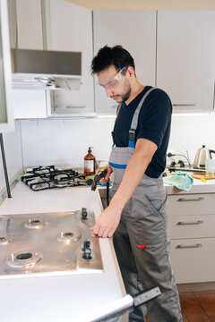 Professional handyman wearing coveralls and safety goggles working on gas hob, performing home appliance repair and maintenance services in modern kitchen..