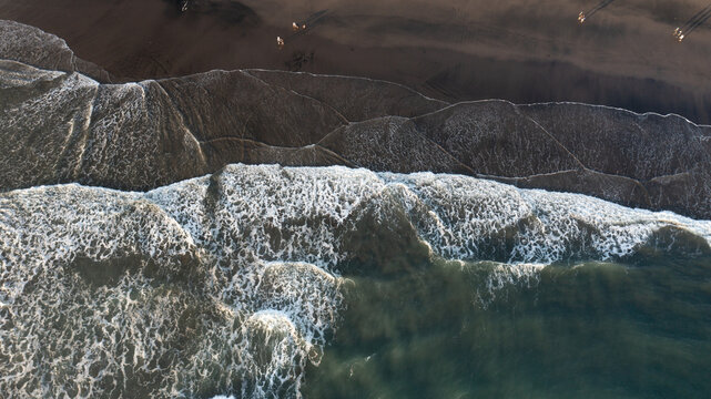 Aerial view of dark volcanic sand beach with crashing white waves and people walking along the shoreline in Piha, Auckland, New Zealand.