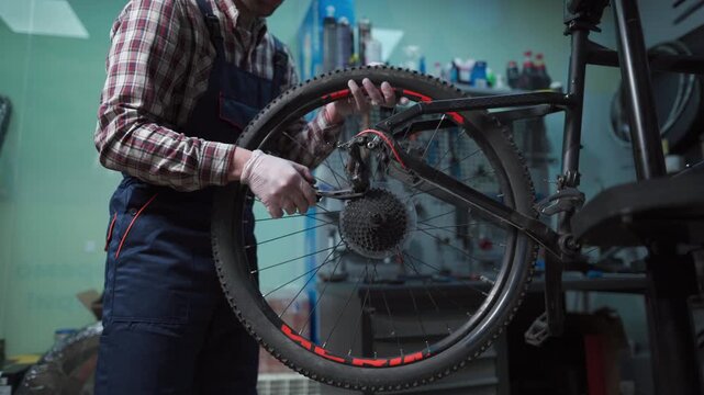 Male mechanic working in bicycle repair shop, mechanic repairing bike using special tool, wearing protective gloves. Young attractive serviceman fixing customer's bicycle wheel at his own workshop.