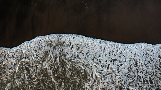 Aerial view of the dark volcanic sand of Piha Beach being washed by white sea foam and crashing waves in Piha, Auckland, New Zealand.