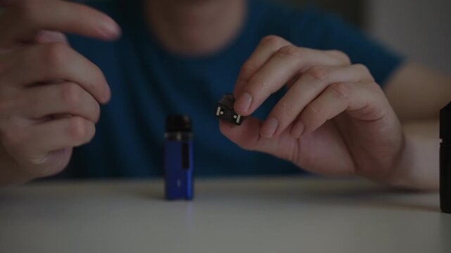 Unrecognizable man in blue t shirt sitting at table unboxing and assembling new liquid cartridge for electronic cigarette, preparing to vape and replacing old part. Shooting in slow motion