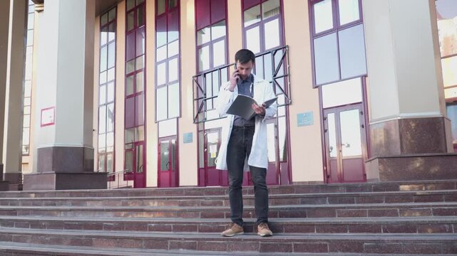 Healthcare professional makes phone call while reading patient chart near hospital. Young male doctor holding a folder with papers and talking on the phone. Telemedicine, telehealth concept.