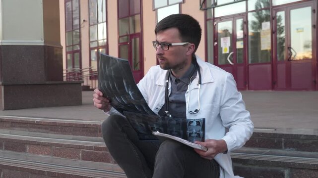Tense doctor man examines x-ray on stairs outside clinic. Medical professional tired and upset checking CT scan of patient in hospital during work shift. Exhausted radiologist looking mri scan chest.